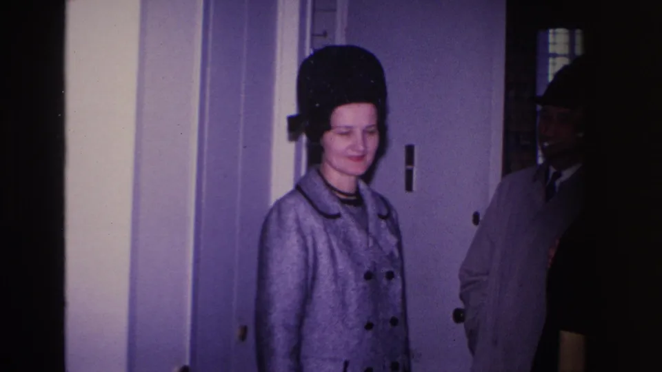 1966 home movie frame: woman in grey double-breasted coat and structured black hat looking down at a white interior door, man in dark suit behind her.