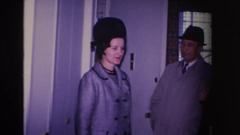 1966 home movie: woman in grey coat and structured black hat beside a man in fedora and light suit at a residential interior doorway, Hoboken, New Jersey.