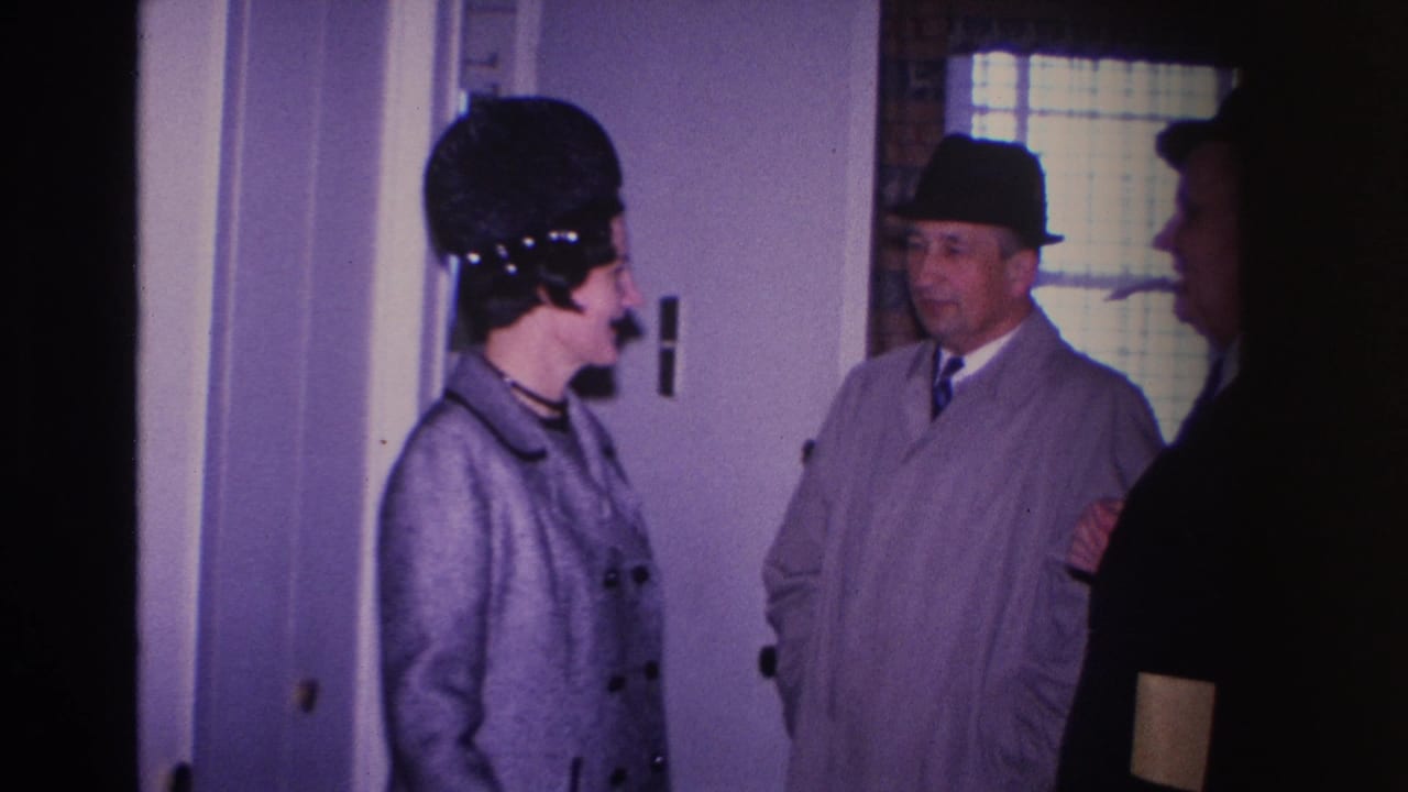 1966 8mm home movie frame — woman in structured black hat and gray coat at an indoor doorway in Hoboken, New Jersey, with a man in a fedora beside her.