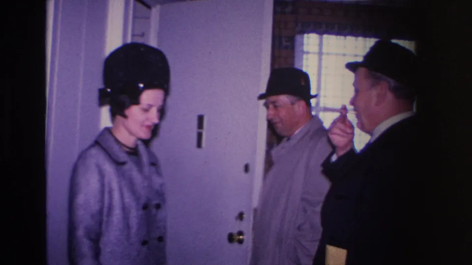 8mm home movie frame from 1966 Hoboken, New Jersey — woman in structured black hat and two men in suits and fedoras near a white interior door; one man holds what appears to be a cigarette.
