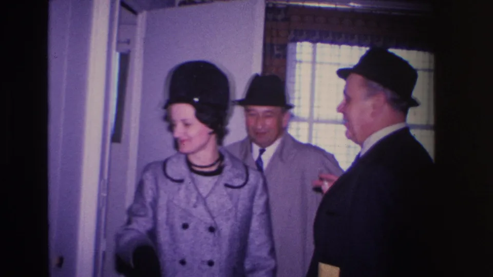 8mm home movie frame from 1966 Hoboken, New Jersey — woman in black hat smiling toward the camera, two men in suits and brimmed fedoras beside her in an interior hallway.