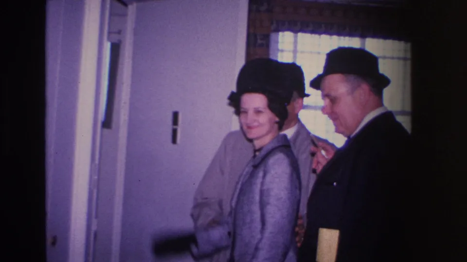 8mm home movie frame from 1966 Hoboken, New Jersey — woman in structured black hat smiling beside a man in a suit and fedora near an interior door, window with blinds in background.