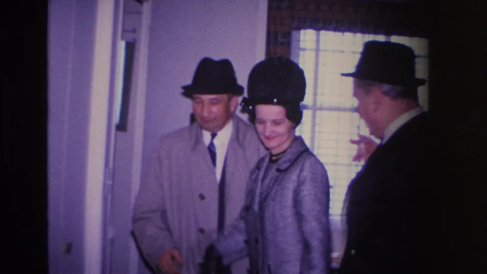 Three adults in mid-1960s dress entering a residential hallway in what is described as Hoboken, New Jersey, 1966; the woman wears a gray coat and black pillbox hat and is smiling.