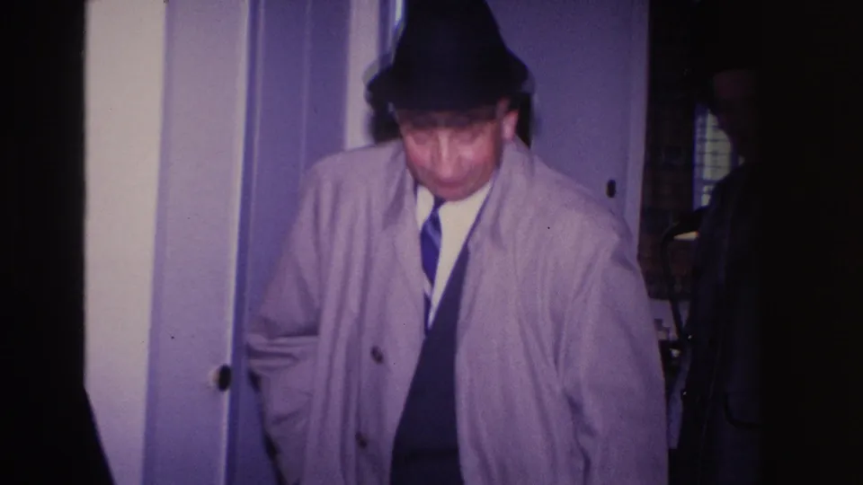Man in a light gray trench coat, dark fedora, and striped tie looking down as he moves through a residential hallway described as Hoboken, New Jersey, 1966.