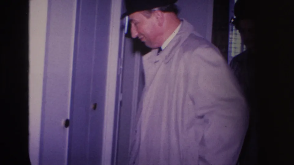Man in a light gray trench coat and dark fedora beside a door frame inside a residence described as Hoboken, New Jersey, 1966; patterned background surface and second figure visible.