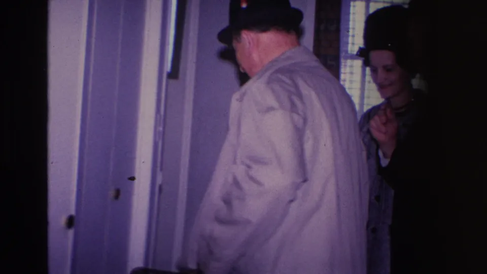 Man in tan trench coat and dark fedora arriving at a home entrance, greeted by a woman in a dark coat and hat — Hoboken, New Jersey, circa 1966.