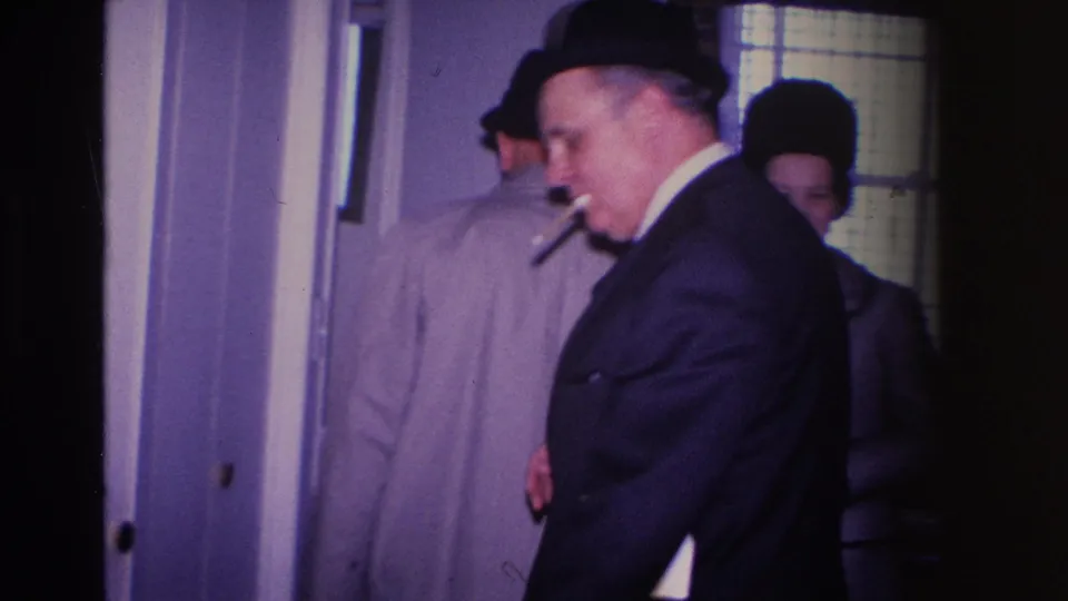 Man in tan overcoat with cigarette, partially turned at a home doorway, other guests visible in background — domestic interior, Hoboken, New Jersey, circa 1966.