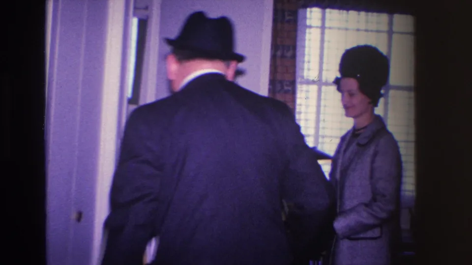 Man in dark suit and hat seen from behind, greeting a woman in a gray coat and fur-trimmed hat in a residential hallway — circa 1966.