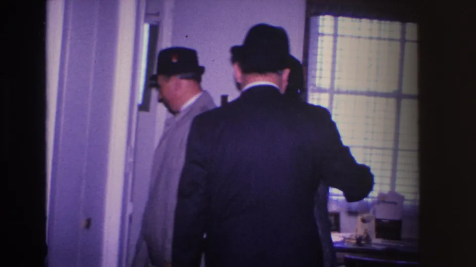 Three adults in mid-century hats and coats congregating in a home entryway, tableware on a surface in the foreground — Hoboken, New Jersey, circa 1966.