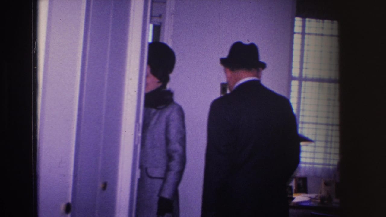 Man in dark suit and fedora stands at interior doorway with woman in light coat and pillbox hat beside him, both viewed from behind.