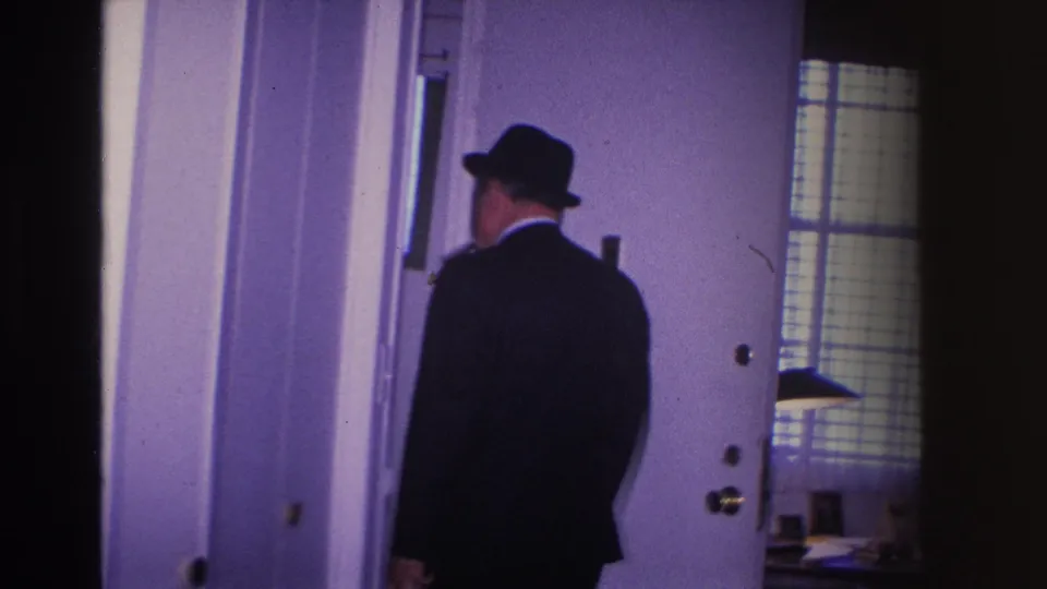 8mm frame from 1966 Hoboken home movie showing a man in dark suit and fedora entering a room through an interior door, woman no longer in frame.