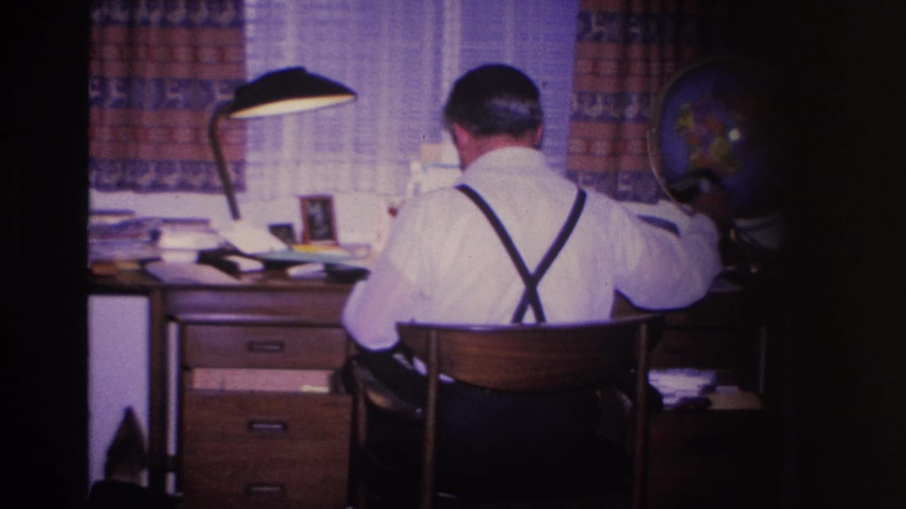 Man in white shirt and black suspenders at a wooden filing desk with flat-shade lamp and decorative globe; 8mm home movie, Hoboken, New Jersey, 1966.