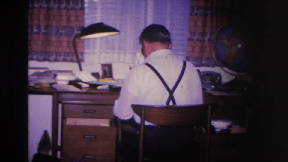 Sustained shot of man in suspenders at mid-century wooden desk; lamp and globe in background; 8mm home movie, Hoboken, New Jersey, circa 1966.