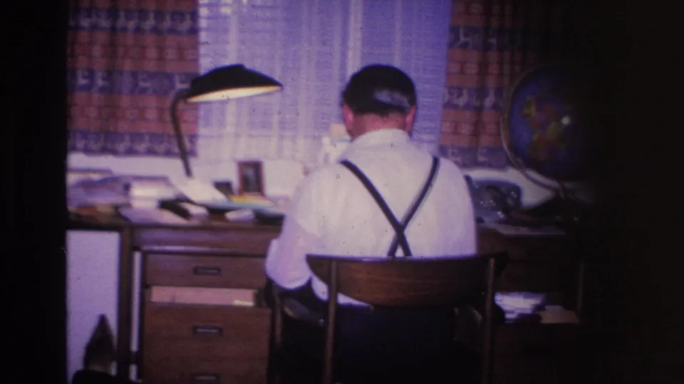 Held frame of man at desk in 8mm home movie; desk lamp, globe, and sheer curtains frame scene; Hoboken, New Jersey, 1966 domestic interior.