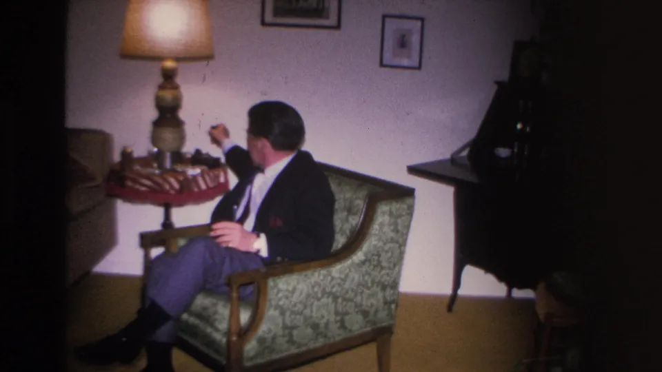 Second man in dark suit and tie in a patterned upholstered armchair; table lamp and framed pictures on white wall behind; 8mm home movie, Hoboken, New Jersey, 1966.