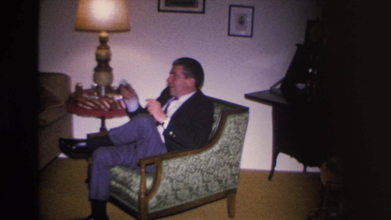 Man in dark suit seated in a mid-century green armchair holding a pipe; decorative lamp and framed wall art visible; 8mm home movie, Hoboken NJ, circa 1966.