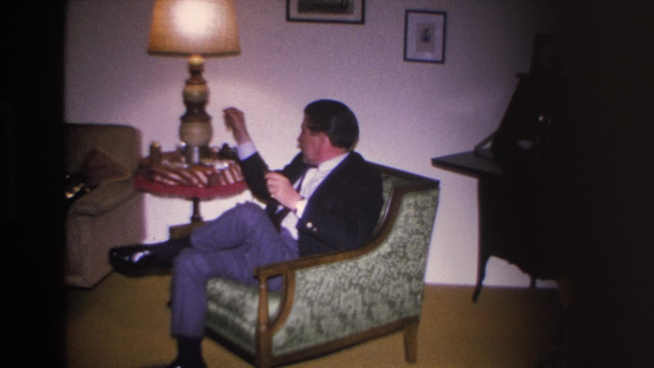 Man in mid-century living room raises hand, likely lighting a pipe; decorative lamp and what appears to be a piano at far right; 8mm footage, Hoboken NJ, circa 1966.