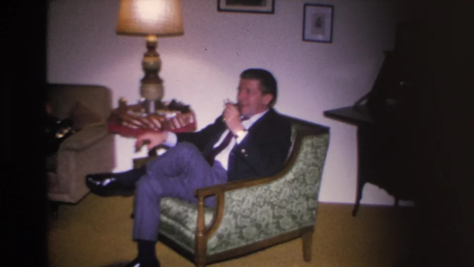 Man draws on a pipe while seated in a mid-century upholstered armchair; decorative table lamp and side table visible; 8mm home movie, Hoboken NJ, circa 1966.