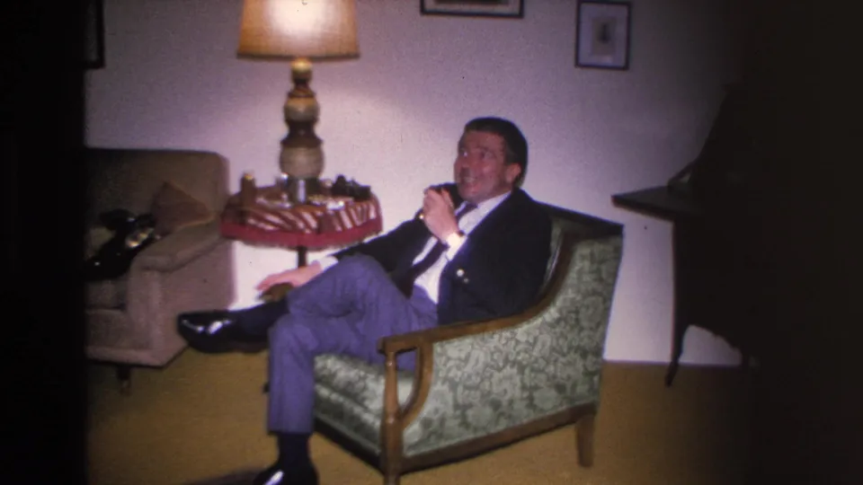Man smoking a pipe in a mid-century living room armchair; table lamp, decorative side table, and partial piano edge at right visible; 8mm footage, Hoboken NJ, circa 1966.
