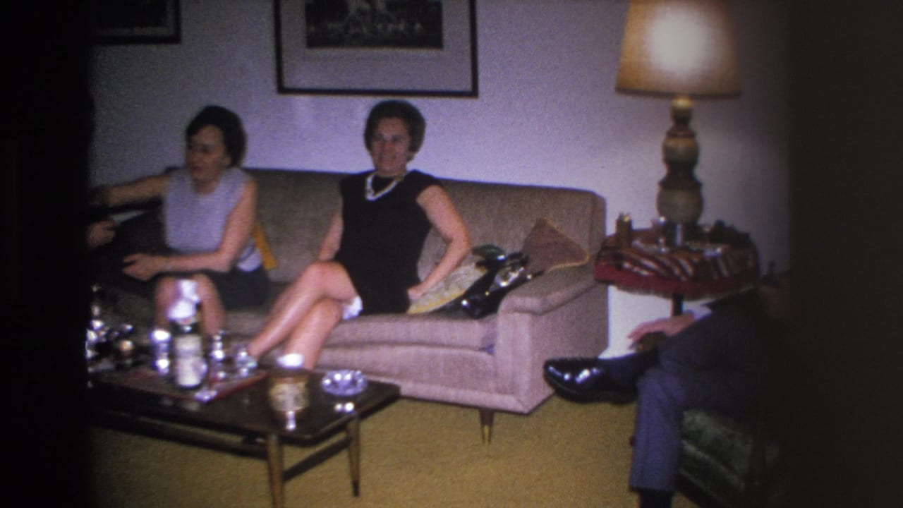 Two women in 1960s shift dresses on a beige sofa in a mid-century American living room, circa 1966; silver tea service and gold carpet visible.