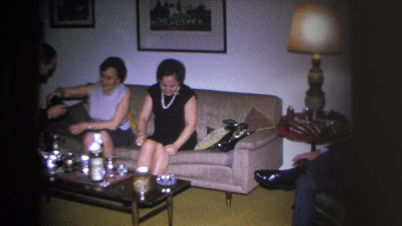 Two women on a beige sofa with a man's dark-suited figure partially entering frame at right; silver tea service on coffee table.