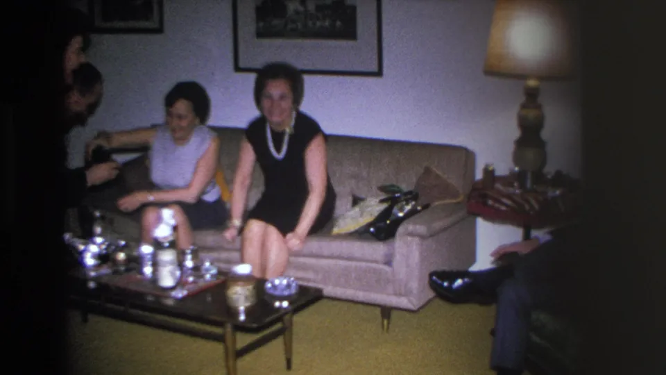 Two women in 1960s sleeveless dresses on a beige sofa in a mid-century American living room; partial figure at left edge; tea service and gold carpet in foreground.