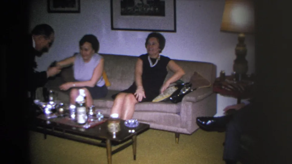 A man's arm reaches toward two women in 1960s shift dresses on a beige sofa in a mid-century American living room; silver tea service on low coffee table in foreground.