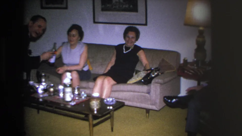 Two women on a mid-century sofa in a 1966 Hoboken home; one in a dark dress with pearl necklace, the other in a sleeveless top; drinks on coffee table.