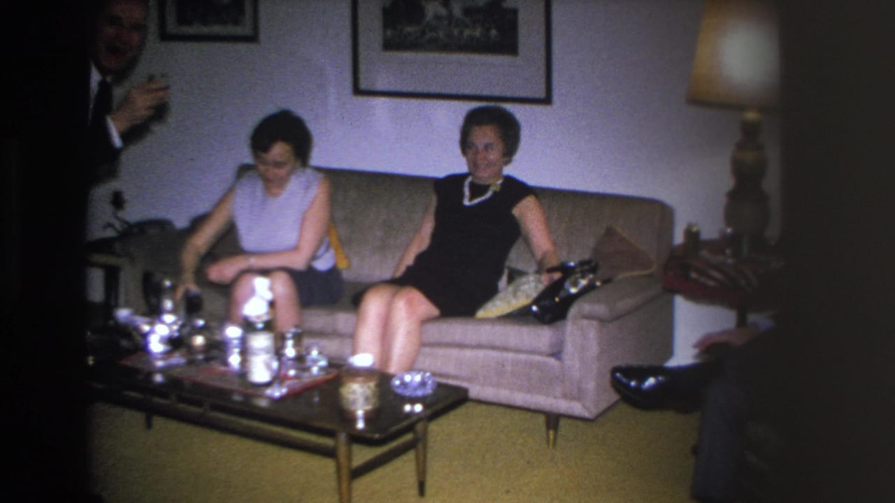 Two women seated on sofa; man at right edge gestures toward them; glassware and bottles on coffee table in foreground.