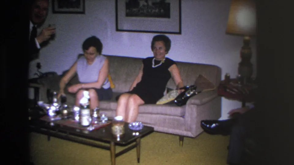 Man in dark suit gestures near two seated women in a 1966 Hoboken living room, drinks set out on a mid-century coffee table.