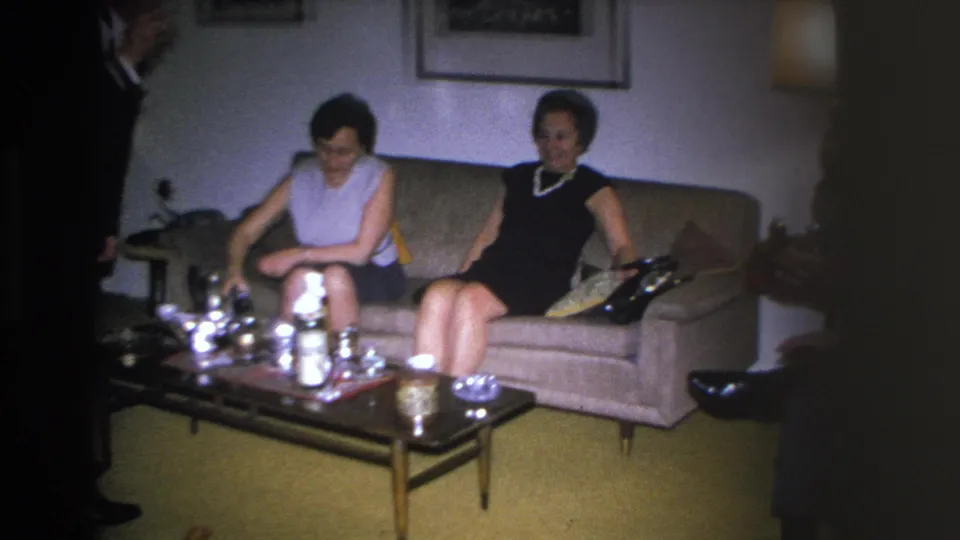 Continuing scene of two women on a mid-century sofa in a 1966 Hoboken living room; coffee table with glassware prominent in foreground.