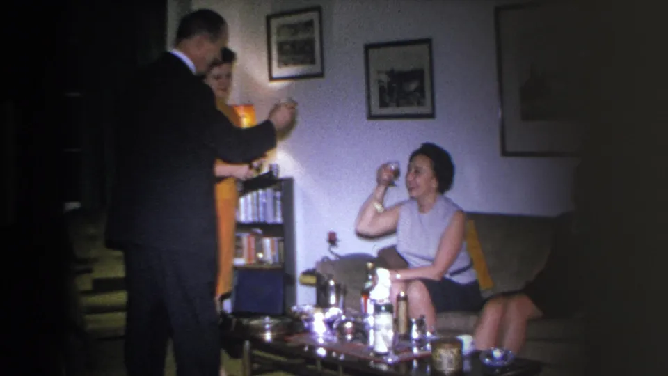 Man serving a drink to a seated woman in a 1966 Hoboken living room; a bookcase stocked with books and two framed prints visible on the right wall.