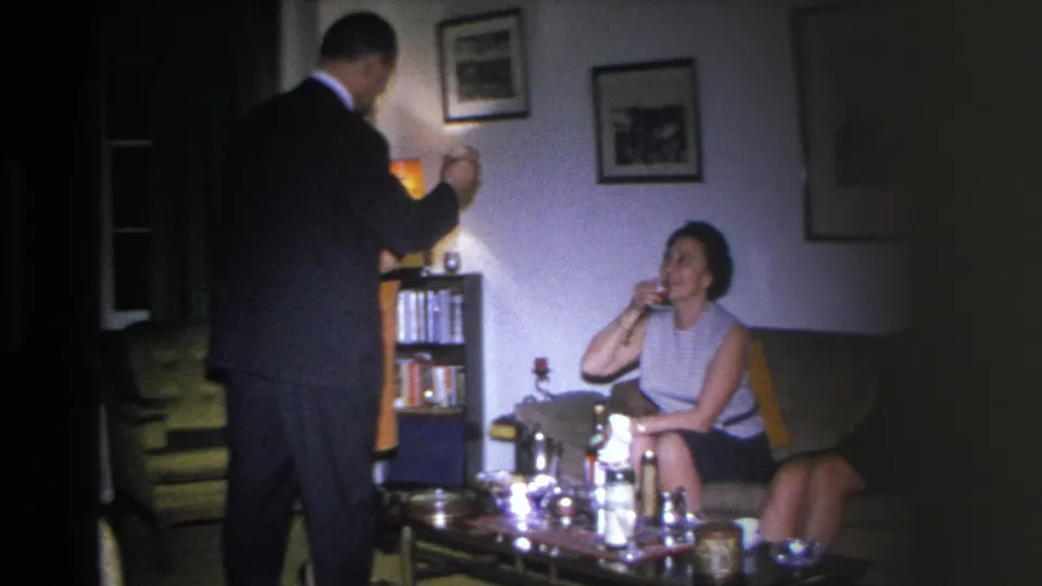 8mm home movie c.1966, Hoboken NJ; man in dark suit standing at left, woman in sleeveless dress seated sipping a drink, mid-century coffee table with bottles in foreground.