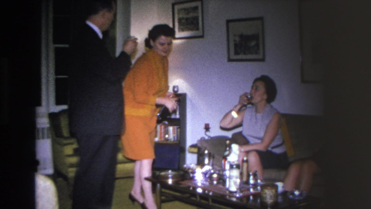 Man in dark suit at left, woman in bright orange dress standing in center, woman in sleeveless top seated at right holding a glass, coffee table with bottles in foreground.