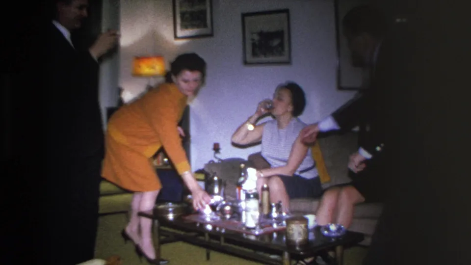 8mm home movie c.1966, Hoboken NJ; woman in orange dress leans toward drinks table, woman in sleeveless top seated on sofa, amber lamp at left, framed pictures on wall behind.