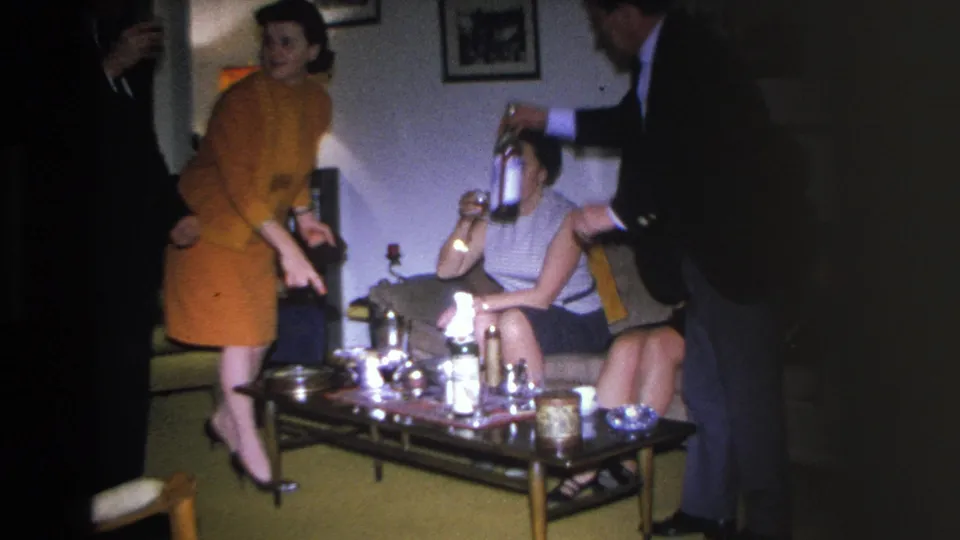 8mm home movie c.1966, Hoboken NJ; woman in orange dress and woman in sleeveless top both seated near coffee table with bottles and glassware, man in dark suit at right edge.