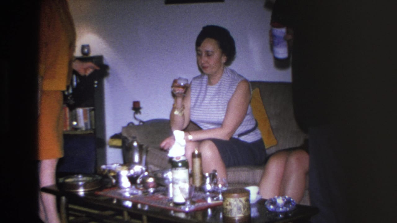 1966 Hoboken home movie: woman on couch holding drink at mid-century living room party, coffee table with bottles visible.