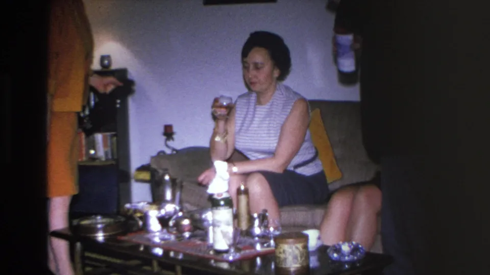 1966 Hoboken home movie: woman on couch holding drink at mid-century living room party, coffee table with bottles visible.