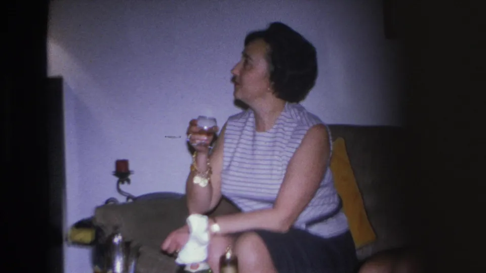 1966 Hoboken home movie: profile close-up of woman in striped top holding drink at living room party, gold bracelet visible.