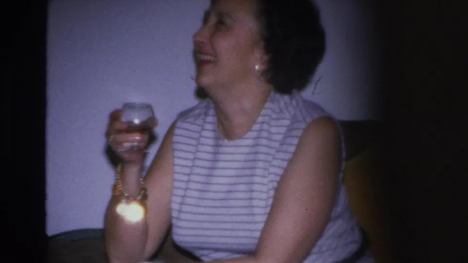 1966 Hoboken home movie: close-up of laughing woman raising a glass at a living room party, gold jewelry visible.