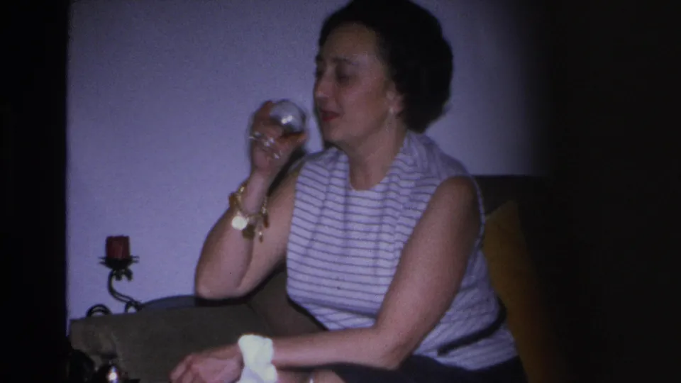 8mm frame of a woman in a 1960s striped sleeveless top raising a glass to her lips; gold jewelry visible, wall sconce and mustard sofa arm in background.