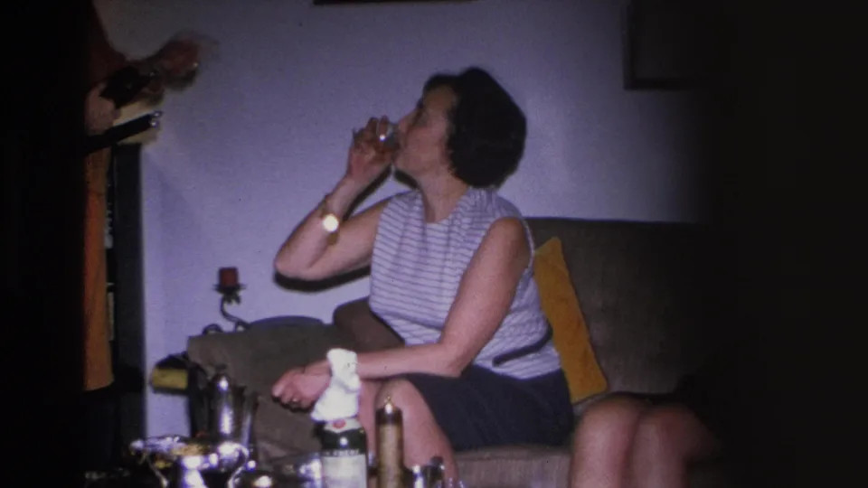 Wide 8mm shot of a woman in 1960s striped top drinking on a couch; coffee table with bottles and cups in foreground, second person partly visible at left.