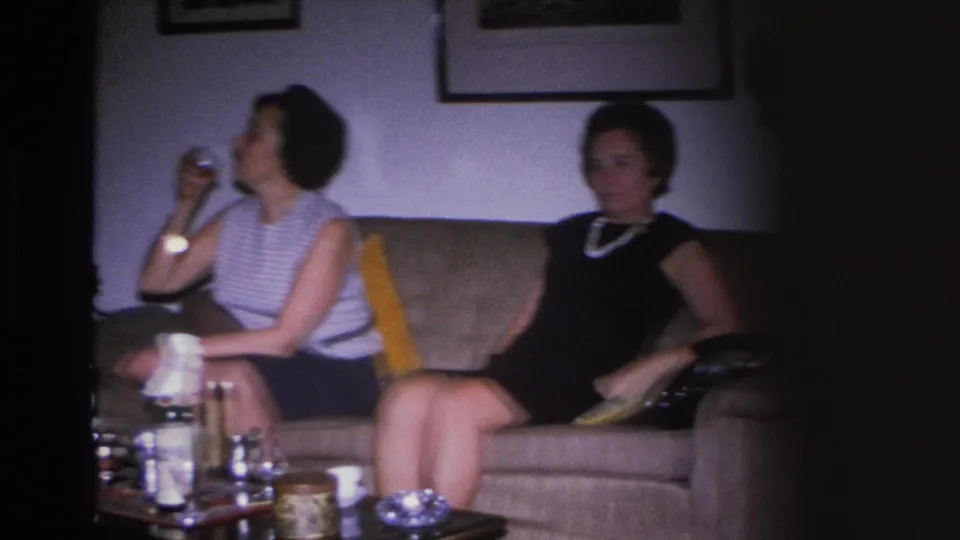 8mm frame of two women on a couch in a 1960s living room — striped top with glass at left, dark dress with pearl necklace at right; bottles and cups on coffee table.