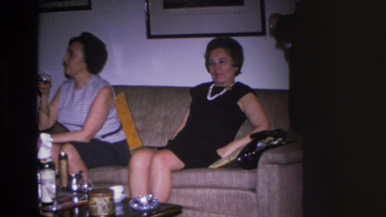 Two women in 1960s attire, one in a black dress with pearl necklace and one in a striped sleeveless top, seated on a tan sofa in a Hoboken home, circa 1966.