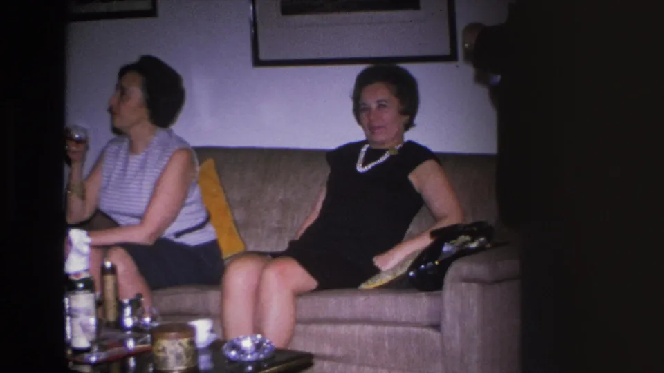 Two women in 1960s attire, one in a black dress with pearl necklace and one in a striped sleeveless top, seated on a tan sofa in a Hoboken home, circa 1966.