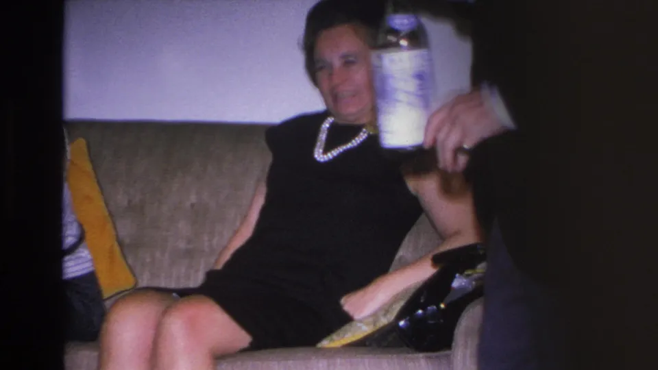 Close shot of a woman in a black shift dress and pearl necklace holding a drink at a home gathering, with a vermouth bottle behind her, Hoboken, circa 1966.