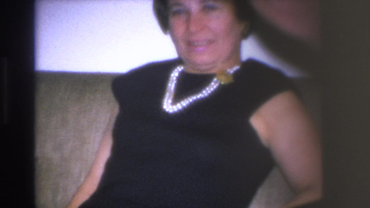 Woman in black cap-sleeve dress and pearl necklace seated on a beige upholstered couch, smiling at camera.