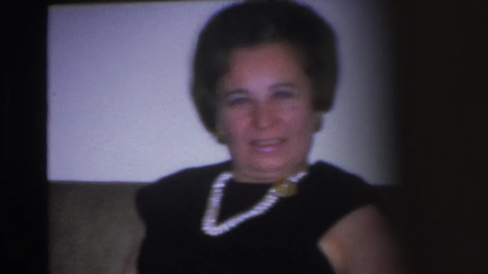 Close-up portrait from a 1966 8mm home movie showing a woman smiling; multi-strand pearl necklace with gold clasp and black dress clearly visible; film grain evident.