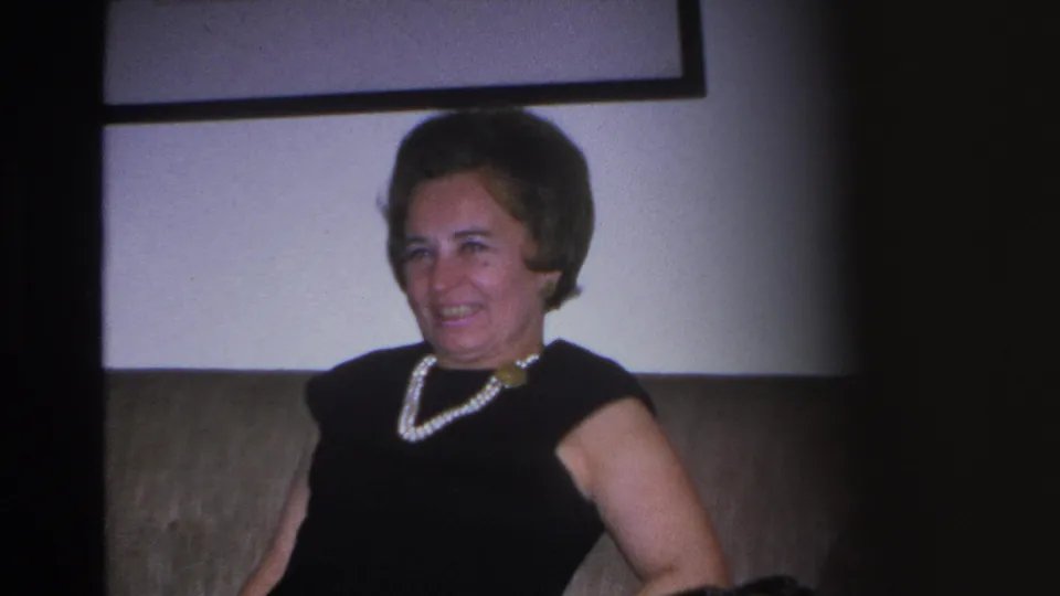 Final frame of a 1966 Hoboken 8mm home movie showing a woman seated on a couch with a framed picture on the wall; pearl necklace with gold clasp and 1960s styling clearly visible.