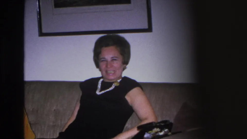 Close-up of a woman in a black dress and pearl necklace laughing on a sofa in a mid-century American living room, circa 1966, Hoboken, NJ.